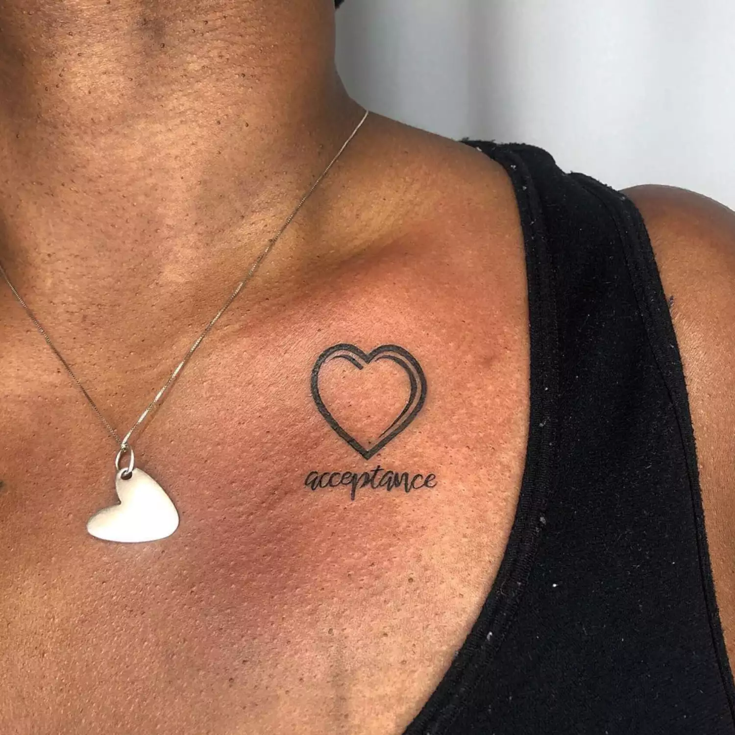 A cartoon heart with the word "acceptance" written in script below it. The tattoo is on the side of the clavicle.