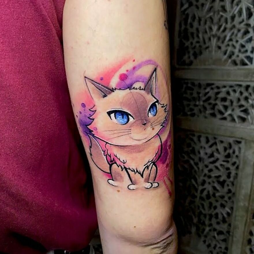 Watercolor tattoo of a cat with blue eyes on an arm, featuring pink and purple hues.