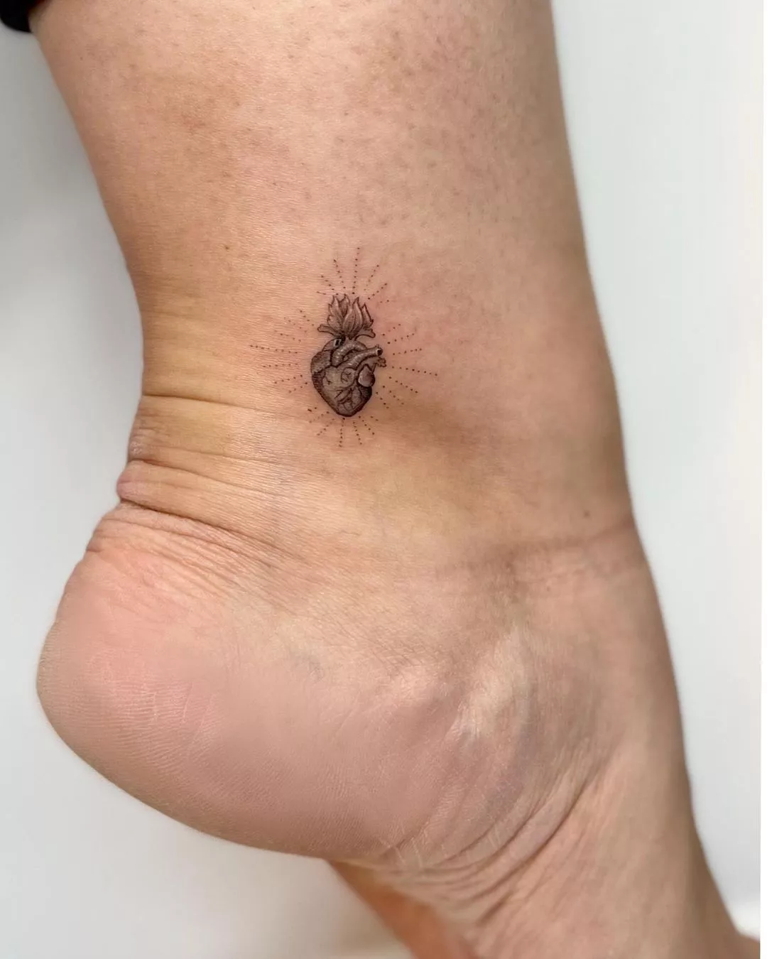 Person shows off the tattoo on their ankle of a sacred heart with linear dots coming out of it
